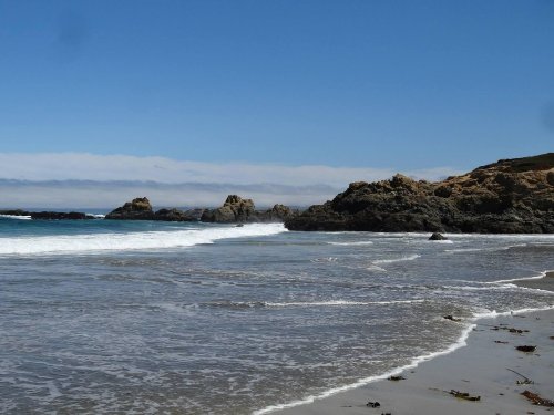 A serene beach scene with gentle waves, rocky formations, and a clear blue sky in the background.