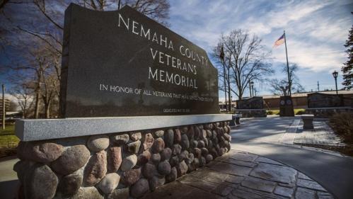 A stone monument for the Nemaha County Veterans Memorial, honoring all veterans, with trees and a flag in the background.