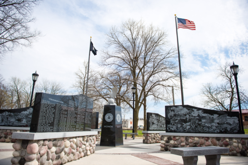 A memorial park featuring stone monuments, flags, and trees against a cloudy sky, honoring veterans and their service.