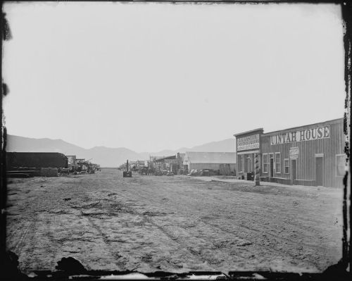 A dusty street lined with buildings, including the Uintah House, with mountains in the background.