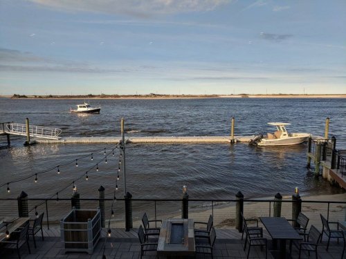View of a river with two boats near a dock, surrounded by outdoor seating and a clear sky.