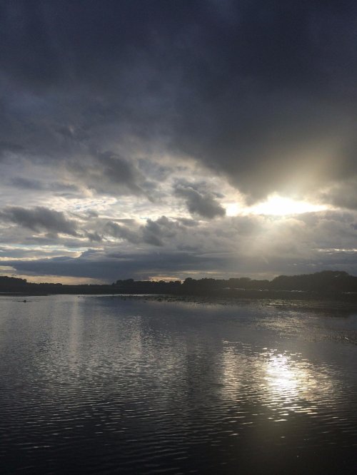 A serene river scene under a cloudy sky, with sunlight breaking through the clouds and reflecting on the water.