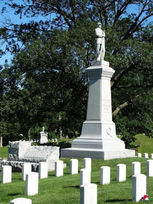 A soldier statue atop a pedestal in a cemetery, surrounded by white gravestones and green trees.