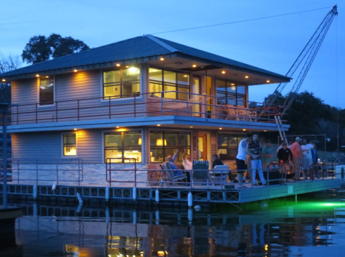 A modern house on a dock at dusk, with people socializing and warm lights glowing from the windows.