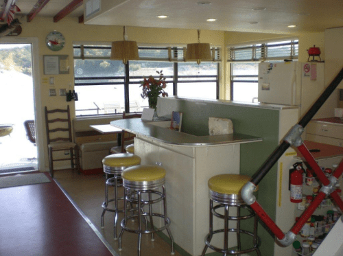 Bright kitchen with a bar, yellow stools, and large windows overlooking a scenic view. Cozy and inviting atmosphere.