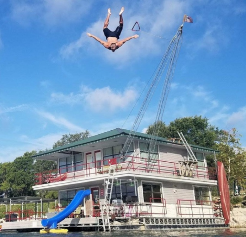 A person dives from a height above a houseboat, with a slide and ladder visible beside the water.