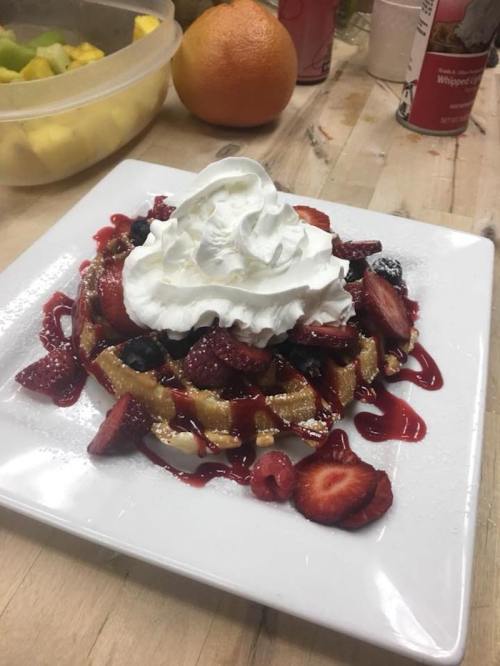 A delicious waffle topped with whipped cream, strawberries, blueberries, and raspberry sauce on a white plate.