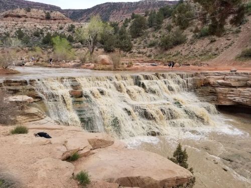 A cascading waterfall flows over rocky terrain, surrounded by greenery and red rock formations in a natural landscape.