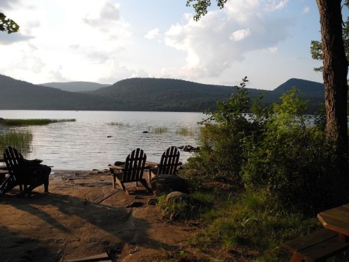 A serene lakeside view with wooden chairs, surrounded by trees and mountains under a partly cloudy sky.