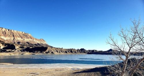 A serene landscape featuring a calm lake, rocky cliffs, and a clear blue sky, with a bare tree in the foreground.