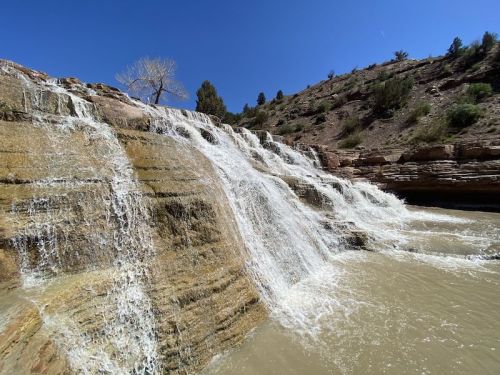 A cascading waterfall flows over rocky terrain, surrounded by trees and a clear blue sky.
