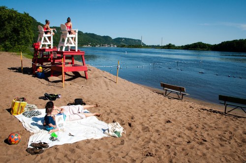 Two lifeguards on chairs oversee a sandy beach by a river, where people relax on towels under a clear blue sky.