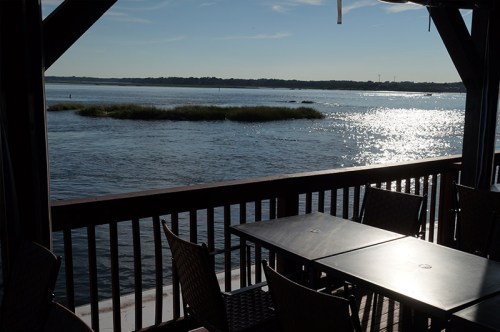A serene view of water and grasslands from a wooden deck, with tables set for dining in the foreground.