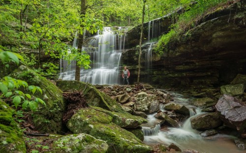 A person stands near a waterfall surrounded by lush greenery and rocky terrain in a serene forest setting.