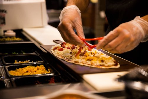 A person in gloves prepares a flatbread with toppings at a food counter, surrounded by colorful ingredients.