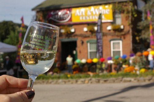 A hand holds a glass of white wine in front of a festival banner and a decorated building with colorful decorations.