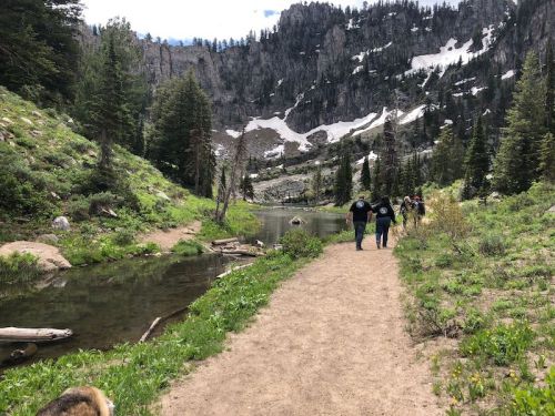 A scenic hiking trail alongside a calm lake, surrounded by mountains and greenery, with people walking in the distance.