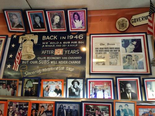 A wall display featuring framed photos, a vintage sign, and newspaper clippings in a sub shop.
