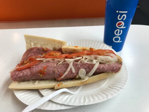 A large sausage sandwich with onions, tomatoes, and sauce, served on a paper plate next to a Pepsi drink.