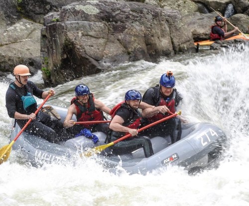 A group of five people in a raft navigating a rapid river, wearing helmets and holding paddles. Water splashes around them.