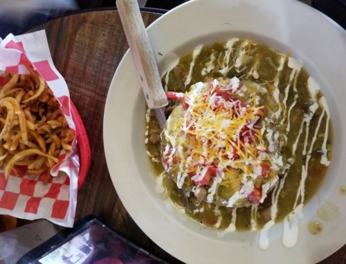 A plate of green chili topped with cheese, tomatoes, and sour cream, alongside a basket of curly fries.