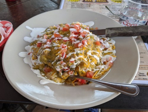 A plate of spaghetti topped with green chili, diced tomatoes, and sour cream, served with a fork and knife.