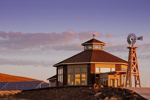 A modern building with large windows, a windmill, and solar panels, set against a colorful sunset sky.