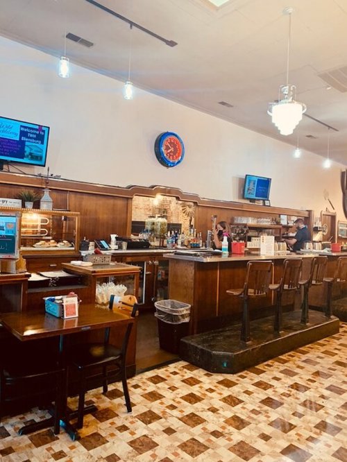 A cozy restaurant interior featuring a bar, tables, and vintage decor, with a clock and screens on the walls.