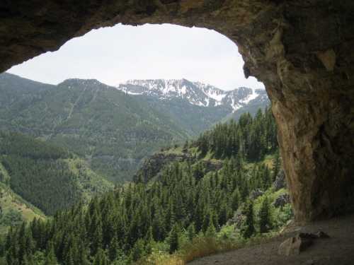 A scenic view from a cave opening, showcasing lush green mountains and snow-capped peaks in the distance.