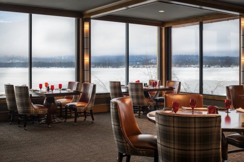Cozy restaurant interior with round tables, leather chairs, and large windows overlooking a snowy lake and mountains.