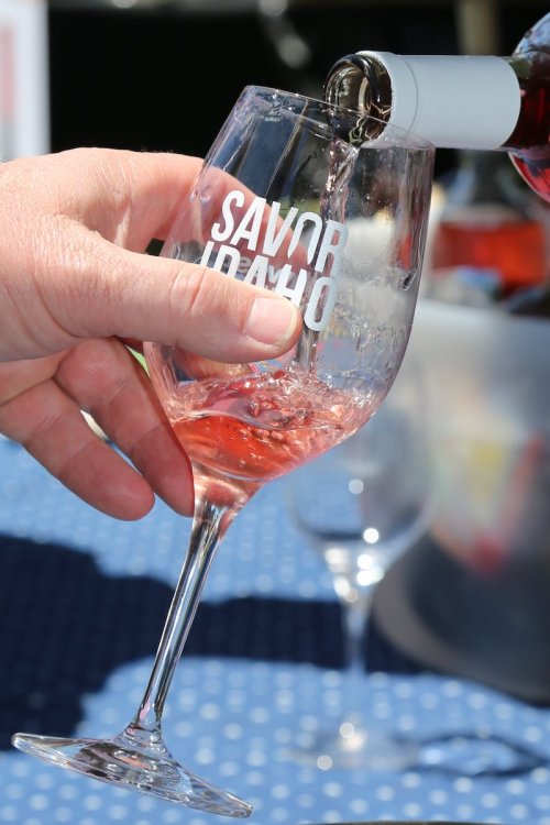 A hand pours rosé wine into a glass with "Savor Idaho" printed on it, set against a blue tablecloth.