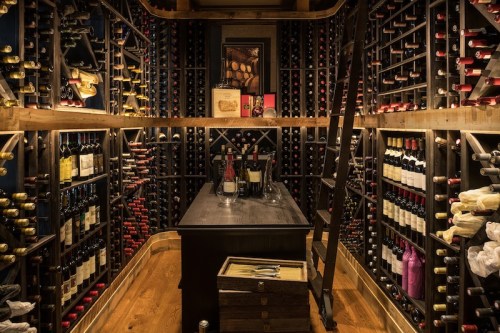 A cozy wine cellar filled with bottles, featuring a wooden table and a ladder against the shelves.