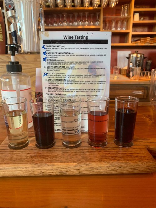 A wooden flight board with five glasses of different wines, alongside a wine tasting menu on a bar counter.