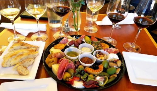 A vibrant charcuterie board with meats, cheeses, olives, and dips, surrounded by glasses of red and white wine.