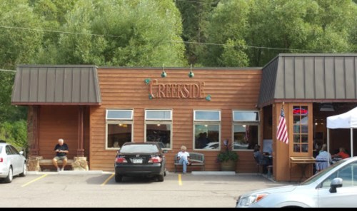 A rustic wooden building with large windows, featuring outdoor seating and an "Open" sign, surrounded by greenery.