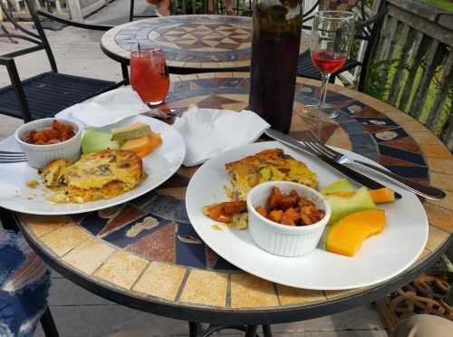 Two plates of food on a mosaic table, featuring frittata, roasted sweet potatoes, and fresh melon, with drinks nearby.