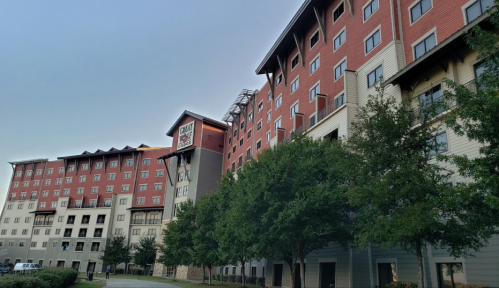 A multi-story building with a mix of red and beige exterior, surrounded by trees and a clear sky.