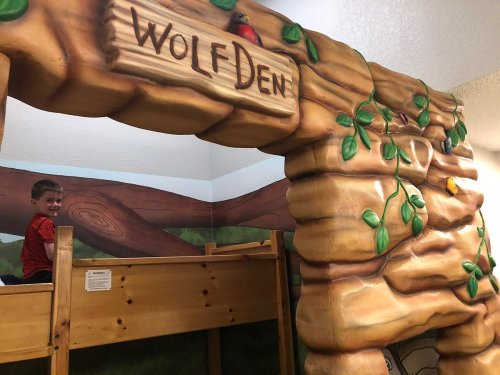 A child smiles while sitting on a wooden bunk bed near a colorful mural of a tree and a sign that reads "Wolf Den."