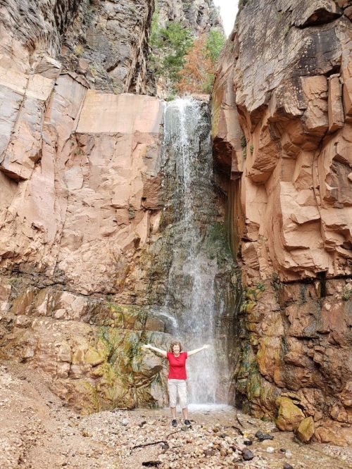 A person stands with arms outstretched in front of a waterfall cascading down rocky cliffs.