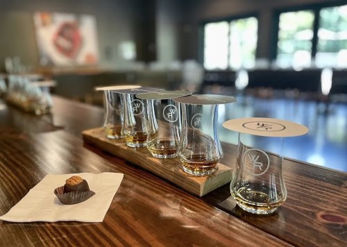 A wooden flight board with five tasting glasses and a chocolate treat on a napkin, set in a modern tasting room.