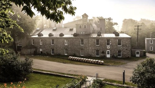 Historic stone building surrounded by greenery, with barrels lined up outside and a misty morning atmosphere.