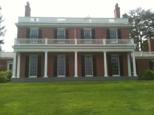 A large brick mansion with white columns, multiple windows, and a balcony, set on a green lawn.