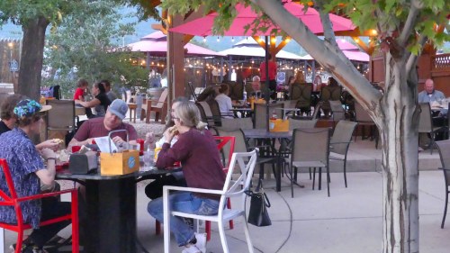 A cozy outdoor dining area with people sitting at tables under umbrellas, surrounded by trees and string lights.