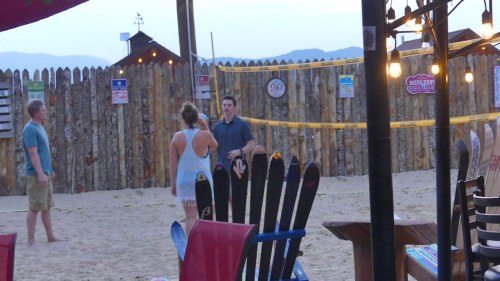 Two people chat on the beach near a volleyball court, with colorful chairs and a rustic fence in the background.