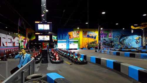 An indoor go-kart track with colorful murals, empty karts lined up, and a digital display in the background.