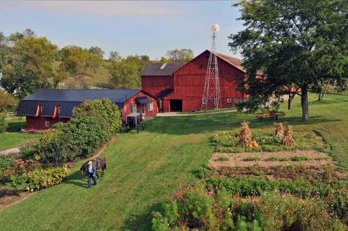 A scenic farm with a red barn, windmill, and a person walking with a cow through lush greenery and gardens.