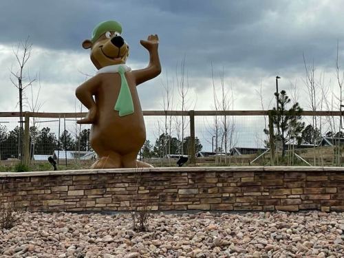 A large bear statue wearing a green hat and tie waves, set against a cloudy sky and rocky landscaping.
