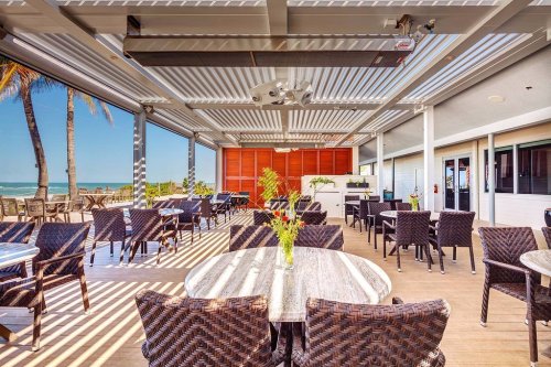 A bright, open restaurant with wicker chairs and tables, featuring large windows and a view of the beach.