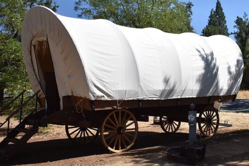 A covered wagon with a white canvas top, resting on wooden wheels in a natural outdoor setting.