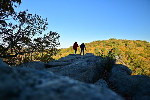 Two hikers silhouetted against a clear blue sky, walking along a rocky outcrop with colorful autumn foliage in the background.